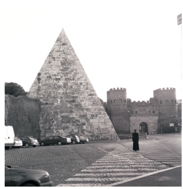A black-and-white photographic portrayal of the same subject as previous images but in the foreground of this Pyramid scene stands a human figure in black, small in contrast to the looming architecture.